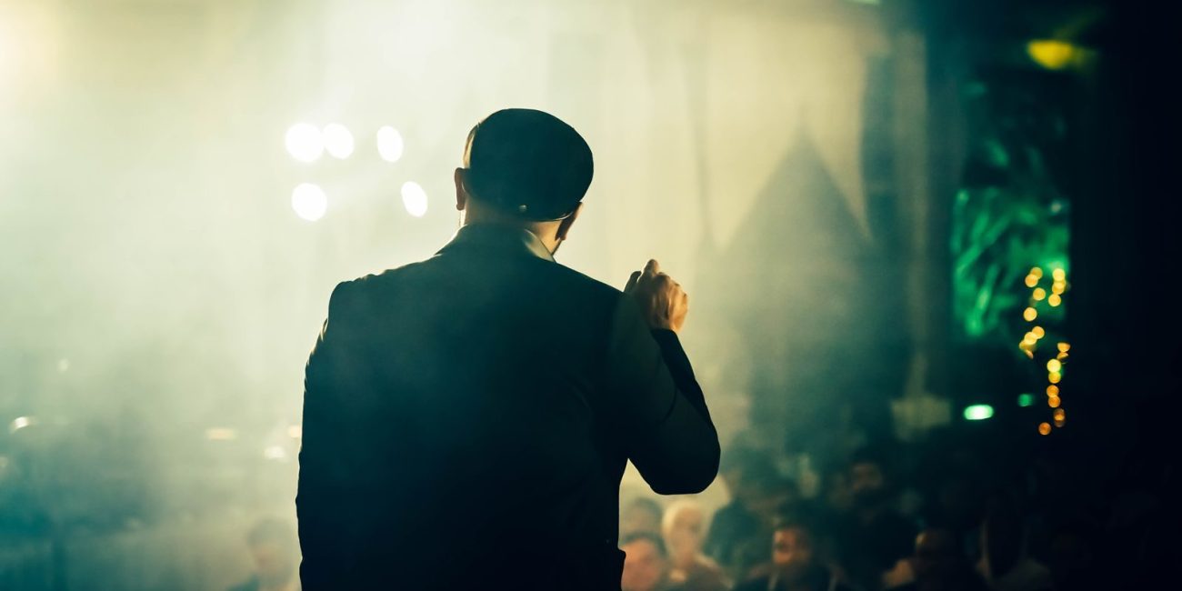 A man seen from the back speaking to an audience at an atmospheric entertainment venue.