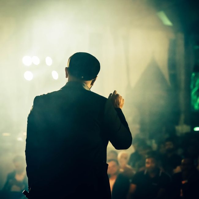 A man seen from the back speaking to an audience at an atmospheric entertainment venue.