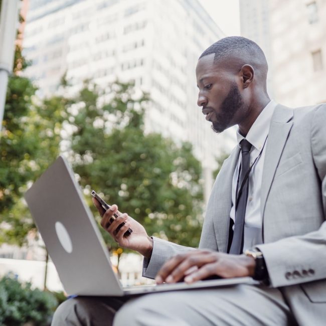 Businessman in a suit using a laptop and smartphone in an urban setting, blending technology with business.
