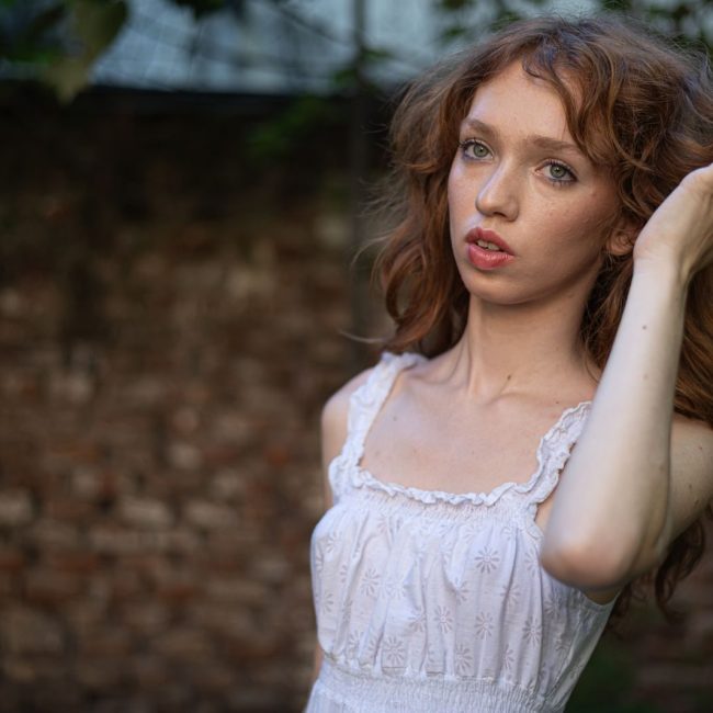 Close-up portrait of a young woman with red hair in a white dress outdoors.