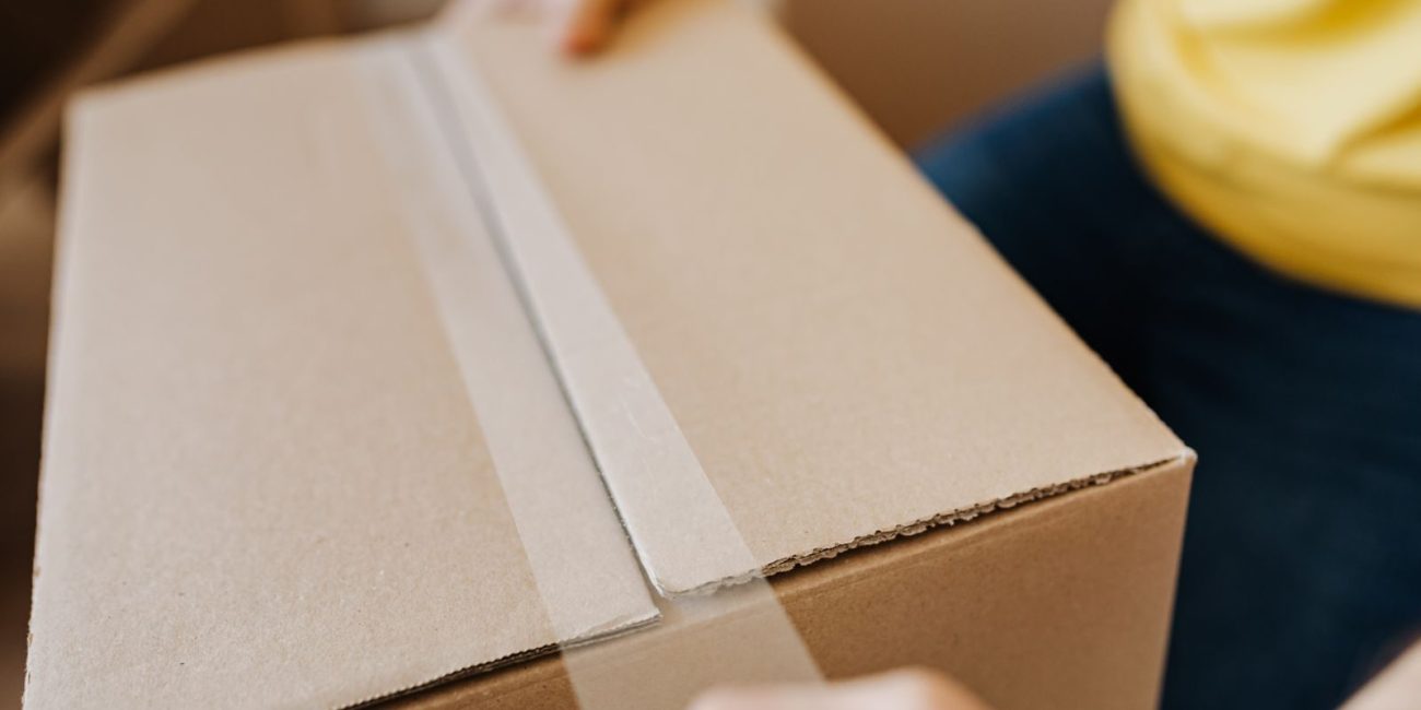 Crop anonymous courier in yellow shirt packing purchase and sealing carton box with tape before deliver