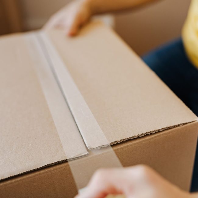 Crop anonymous courier in yellow shirt packing purchase and sealing carton box with tape before deliver